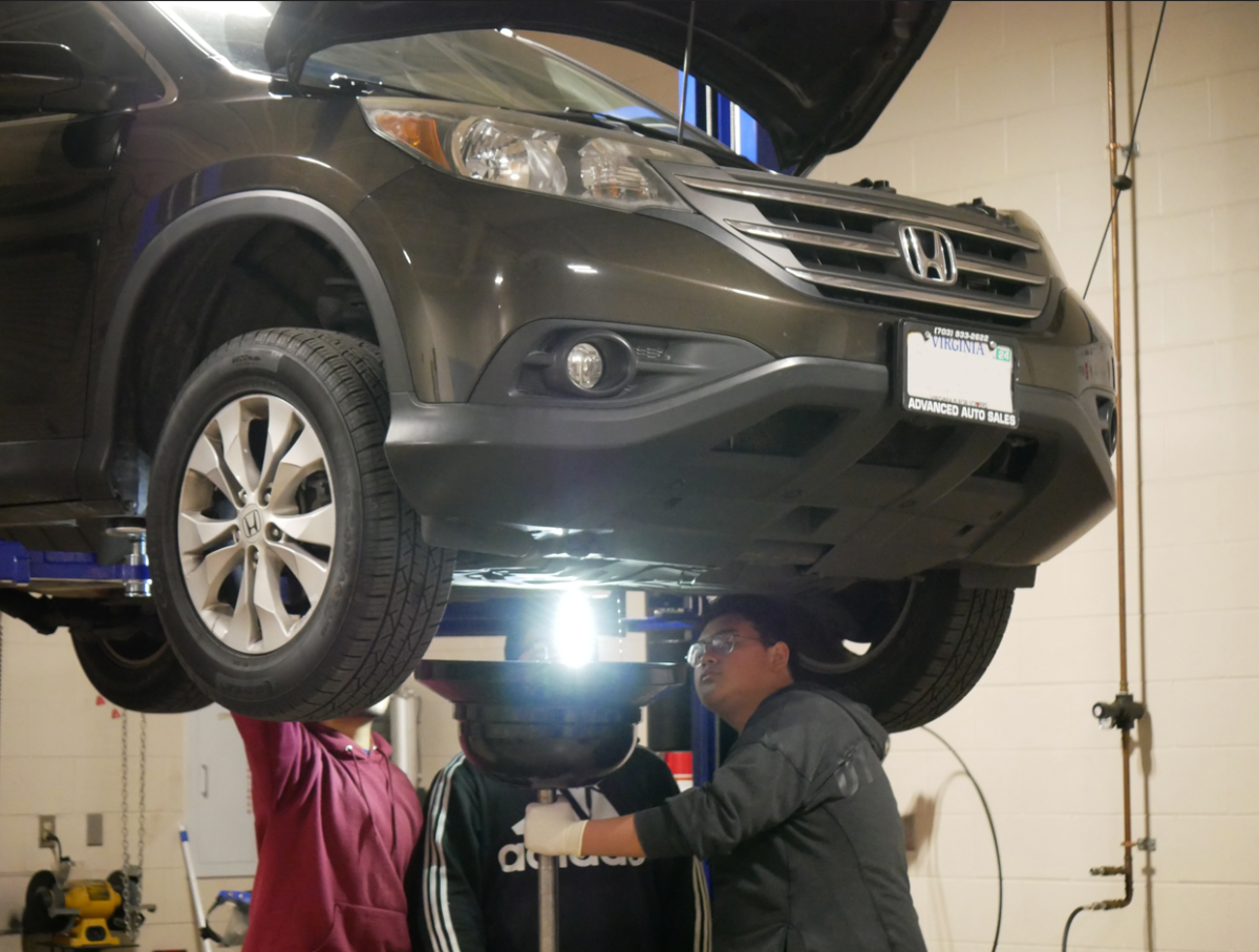 Three students work on a car on November 29 afterschool. Three students perform an oil change on a car, which is suspended from the ceiling with the hood open. Wearing safety goggles, one holds the oil pan. The license plate has been blurred out.
