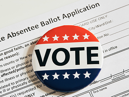 A photograph depicting a pin labeled “VOTE” on a ballot application