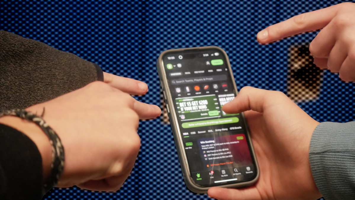 A crowd of students surrounding a phone with a sports betting app emphasizes the increasing number of student gamblers and the ease in which someone can get addicted. 