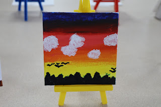 A canvas painted by Eliza Duncan features a sunset with mountains on the horizon and birds flying over them.